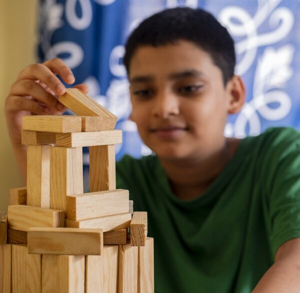 A young boy wearing a green tshirt places a wooden block on a tower made of the same wooden blocks.