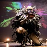 A man bends on one knee. He bald head is covered with paint and he is wearing elaborate feathered clothes.