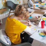 A child sits at a cluttered table full of paints and brushes