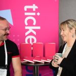 A man and a woman stand either side of a Ticketsolve banner and a table full of bright pink notebooks