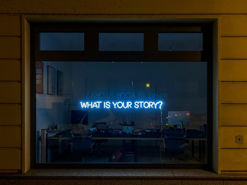 The outside of a window looking into an office. A light spells out: What is your story?