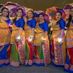 Six women stood together wearing colourful saris and holding parasols. They are each wearing a colourful, glowing wristband.