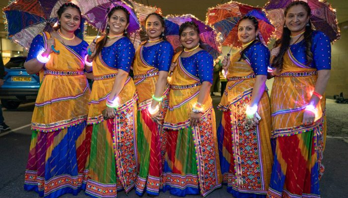 Six women stood together wearing colourful saris and holding parasols. They are each wearing a colourful, glowing wristband.