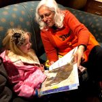 A woman smiles as she shares a large storybook with a small child. The child beams with delight.