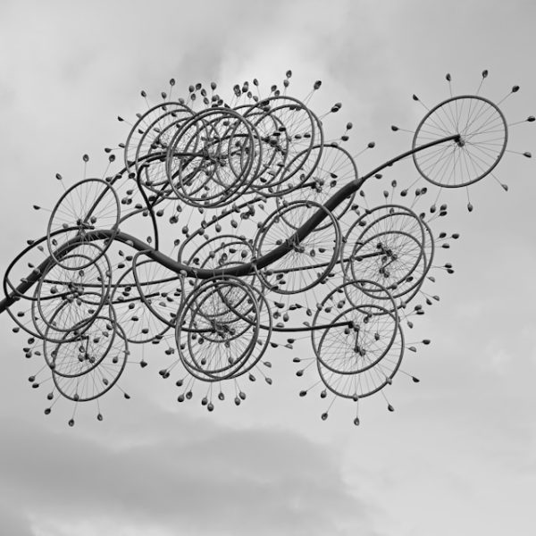 A black and white artistic photograph showing multiple bicycle wheels suspended in the air against a cloudy sky. The wheels are arranged in a chaotic, overlapping cluster, creating an abstract sculpture-like formation. Small decorative elements or lights appear to radiate from the wheels like spokes or stars. The composition creates a dreamlike, surreal effect with approximately 15-20 bicycle wheels of various sizes floating together