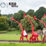 A parade of life-sized, crafted, bright red giraffes against a backdrop of parkland.