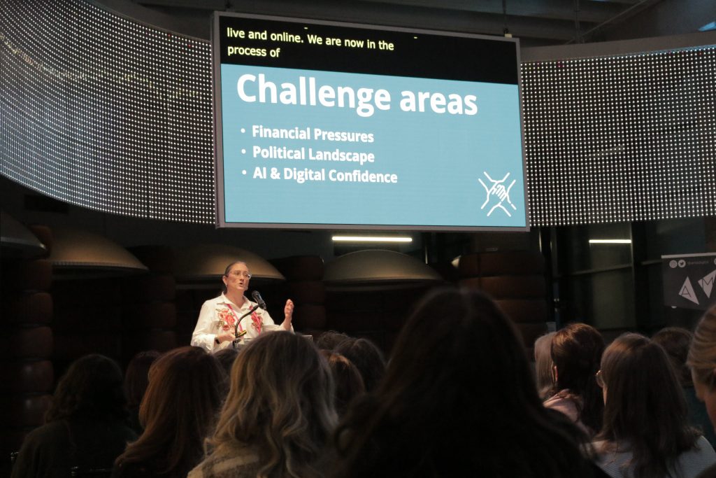 Cath Hume stands at a podium. The screen behind her details 3 challenge areas
