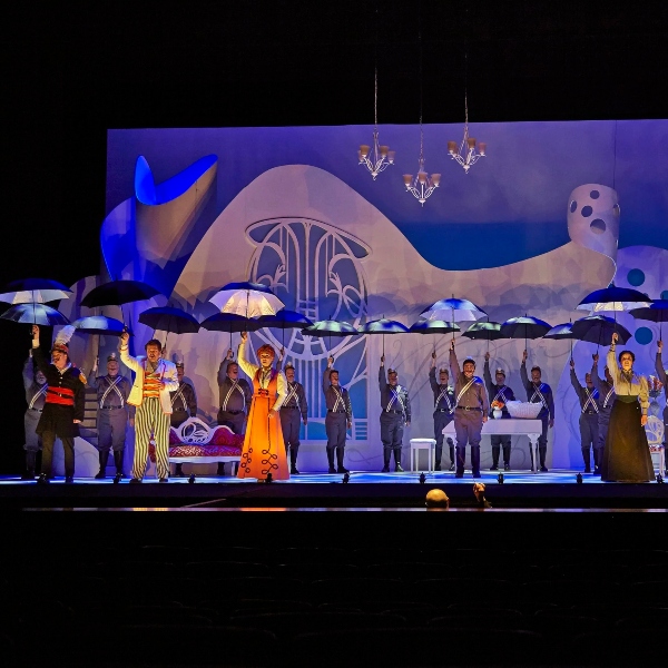 The chorus on stage in costume holding up black umbrellas