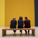 3 people sitting on a bench looking at a floor to ceiling piece of art