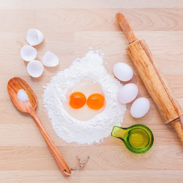 A mound of clour with two cracked egg yolks in the centre with a large wooden spoon to one side and a rolling pin to the other.